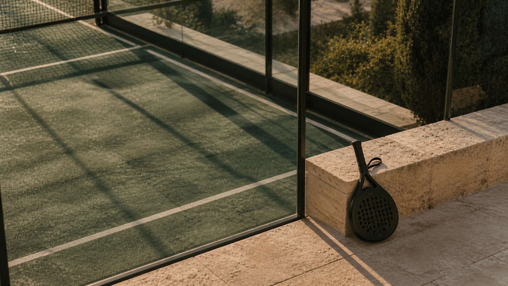 Long shadows crossing a quiet Mediterranean padel court beside a resting racquet